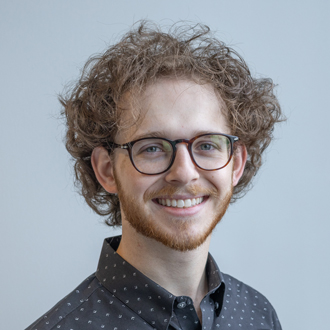 Maxwell facing forward, smiling, curly short hair, glasses, dark shirt