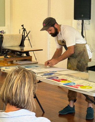 Raymer bending over a table of prints, wearing a white t-shirt, work apron and baseball cap.