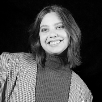 Paige facing forward, smiling, short hair, checkered jacket over a turtleneck sweater