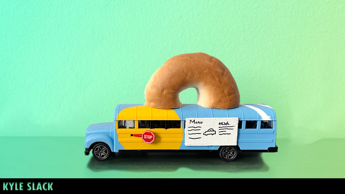 A toy bus with a large bagel atop its roof sits before a green background. The bus is mostly baby blue with a yellow circle design on the front and white striping across the rear of the roof. A menu is posted next to a service window for customers.