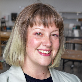 Delaney facing forward, smiling, short blonde and brown hair, beige jacket, black shirt