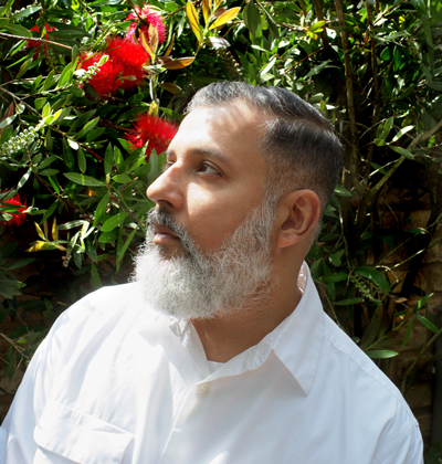 Designer Ashish Gupta in profile looking to his right with a red flower bush behind him.