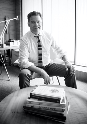 Greg Watts sitting in front of a large window, smiling