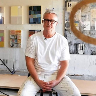 Jim sitting on a table in his art studio with paintings on the wall in the background, wearing glasses, white T-shirt